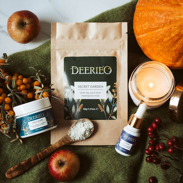 Deerieo Discovery and Travel Set of face cream, facial oil serum and exfoliating facial mask on a green cashmere jumper surrounded by red apples, soy candles and orange pumpkin in Autumnal decoration.