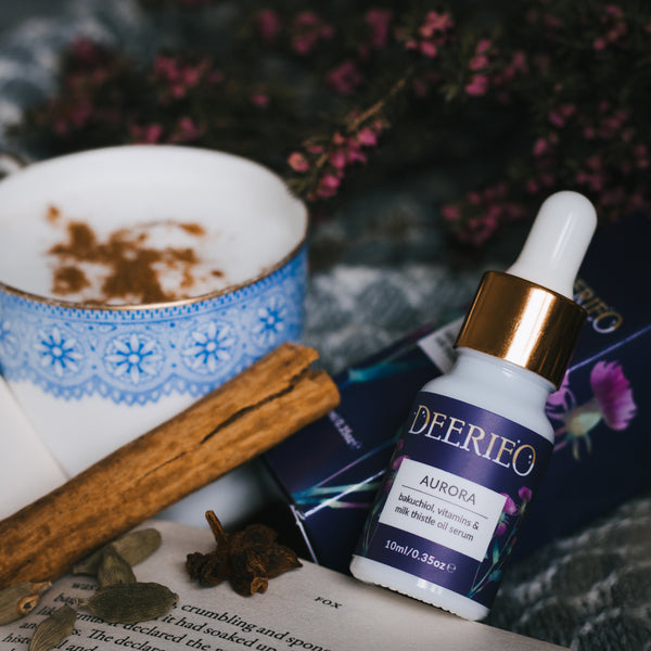 Deerieo Aurora rejuvenating oil serum with bakuchiol, vitamin C and coenzyme Q10 on a grey blanket with open book, cinnamon, spiced latte and purple heather.